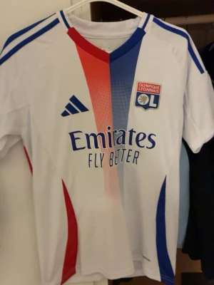 Olympique Lyonnais matchtröja Adidas - Snygg vit matchtröja från Olympique Lyonnais med röda och blå detaljer samt klubbmärke på bröstet. Adidas-logga och 'Emirates Fly Better' tryck på framsidan. Tillverkad i lätt och ventilerande material, perfekt för träning eller match.