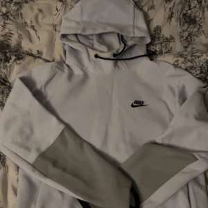 Vit Nike hoodie med grå detaljer - Säljer en snygg vit hoodie från Nike med grå paneler på ärmarna och klassisk logga på bröstet. Hoodien har en stor huva med dragsko och är tillverkad i mjukt material som passar perfekt för en avslappnad streetstyle.