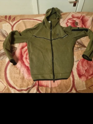 Olivgrön Nike hoodie med dragkedja - Obs!!! Pris går att diskuteras!!! Säljer en olivgrön hoodie från Nike med hel dragkedja och huva. Hoodien har svarta detaljer, en diskret Nike-logga på bröstet och en ficka med dragkedja på ena ärmen. Perfekt för chill dagar eller träning.