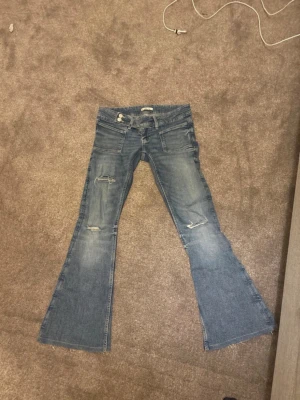 Blå bootcut jeans med slitningar - Snygga blå bootcut jeans med slitningar på benen och klassisk femficksdesign. Jeansen har låg midja och är tillverkade i denim med en lätt tvättad look. Perfekt för dig som gillar en avslappnad och trendig stil.