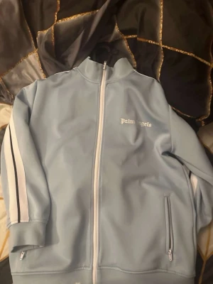 Palm Angels ljusblå zip-tröja - Ljusblå zip-tröja från Palm Angels med vita detaljer och logga på bröstet. Tröjan har höga krage, långa ärmar med vita ränder längs sidorna och två fickor med dragkedja. Perfekt för en sportig och trendig look.