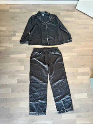 Svart satinpyjamas med vita detaljer - Helt ny Stilren grå pyjamas i satin med långärmad skjorta och byxor. Pyjamasen har vita kontrastsömmar längs krage, ärmslut och benslut. Skjortan har klassisk krage och knäppning framtill, byxorna har resår i midjan för extra komfort.