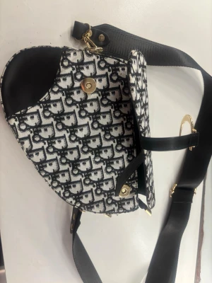 Svartvit axelväska  - Trendig axelväska  i svartvitt mönster med tydlig logga. Väskan har guldfärgade metalldetaljer, bred svart axelrem och detaljer i svart läder. Sadelinspirerad form och magnetlås framtill. Perfekt för dig som vill sticka ut med en ikonisk accessoar.