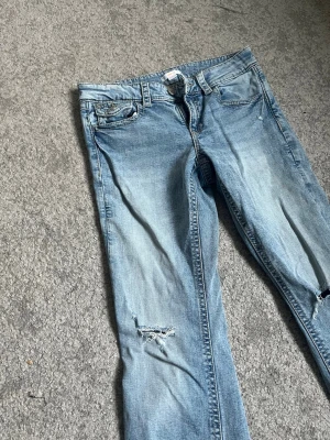Ljusblå slitna jeans från Gina Tricot - Säljer ett par ljusblå jeans från Gina Tricot med slitningar på benen och klassisk femficksmodell. Jeansen har dragkedjegylf och bälteshällor, perfekt för en avslappnad och trendig look.