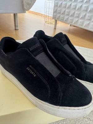 Svarta sneakers från Axel Arigato - Stilrena svarta sneakers från Axel Arigato i mocka med vit sula och diskret logga på sidan. Slip-on modell utan snörning. Väldigt bra skick. 