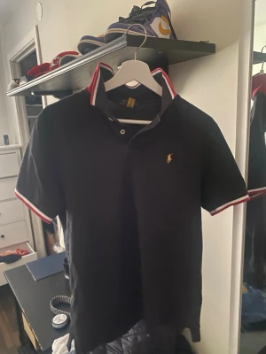 Svart pikétröja från Ralph Lauren - Svart pikétröja från Ralph Lauren med klassisk krage och knappar framtill. Tröjan har röda och vita detaljer på krage och ärmslut samt den ikoniska gula loggan broderad på bröstet. Perfekt för en stilren och sportig look.