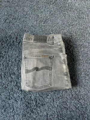 Svarta jeans från Nudie Jeans - Säljer ett par svarta jeans från Nudie Jeans i storlek 32/34. Klassisk femficksmodell med dragkedja och knapp. Jeansen har en rak passform och är tillverkade i denim. Perfekta för dig som gillar stilrena och tidlösa plagg.