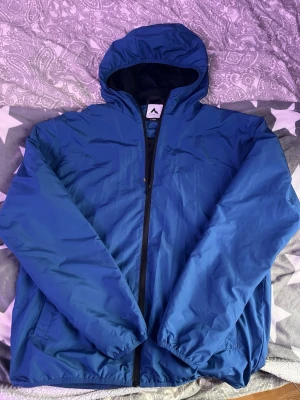 Blå vindjacka med huva - Snygg blå vindjacka från Mountain Ridge med hel dragkedja och praktisk huva. Jackan har två sidofickor och elastiska muddar vid ärmsluten. Materialet är lätt och vindtätt, perfekt för blåsiga dagar. Enkel och stilren design som passar till det mesta.