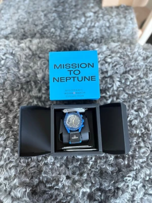 Omega x Swatch Mission to Neptune - Snygg Omega x Swatch Mission to Neptune klocka från Bioceramic MoonSwatch Collection. Klockan har en blå boett, svart urtavla med vita detaljer och svart armband med Omega-logga. Perfekt för dig som gillar unik design och coola färger.