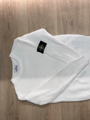 Vit stickad tröja från Stone Island - Snygg vit stickad tröja från Stone Island med klassisk rund halsringning och ribbade muddar. Tröjan har den ikoniska svarta logotypen med kompass på ärmen. Perfekt för dig som gillar stilrena och trendiga plagg.
