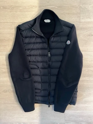 Svart pufferjacka från Moncler - Snygg svart pufferjacka från Moncler med quiltad front och stickade ärmar. Jackan har dragkedja hela vägen, hög krage och Moncler-logga på bröstet. Perfekt för kalla dagar och ger en stilren look.