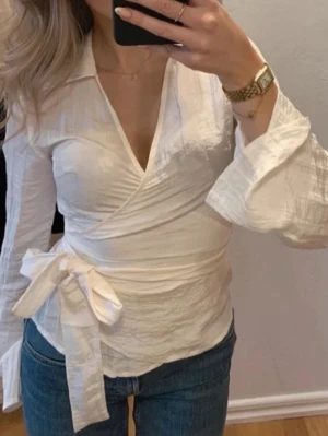 Vit omlottblus med knyt - Snygg vit omlottblus med v-ringning och knyt i sidan. Blusen har långa, lätt utsvängda ärmar. Perfekt för dig som gillar stilrena och trendiga plagg med en feminin touch.