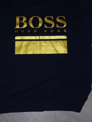 Svart hoodie från Hugo Boss - Svart hoodie från Hugo Boss med stort guldigt tryck på bröstet där det står 'BOSS HUGO BOSS' och en bred guldig rektangel under. Materialet är mjukt och bekvämt, perfekt för en cool och avslappnad look.