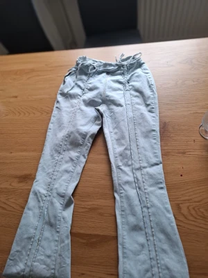 Ljusblå bootcut jeans med dragkedja i midjan - Säljer ett par ljusblå bootcut jeans med raka ben och coola sömdetaljer längs benen. Jeansen har en dragsko i midjan för skön passform och en avslappnad look. Perfekta för dig som gillar en casual stil och vill ha något som sticker ut lite, har använt dom ett par gånger och det är lite trasigt och nertrampat längst nere men annars är dom jättebra.