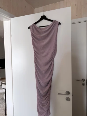 Ljuslila glittrig långklänning - Säljer en snygg ljuslila långklänning med glittrigt tyg och draperad effekt längs hela klänningen. Klänningen är ärmlös och har en elegant, figurnära passform som ger en riktigt trendig look.