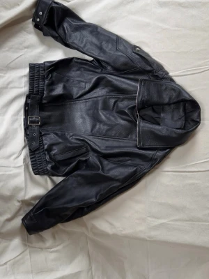 Svart skinnjacka med bälte och hög krage  - Säljer en svart skinnjacka i klassisk bikerstil med bred krage och bälte i midjan. Köpt secondhand, okänt varumärke. 
