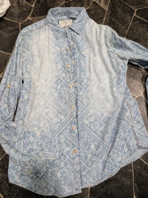 Blå mönstrad skjorta från Denim & Supply stl L - Säljer en blå långärmad skjorta från Denim & Supply med vitt blommigt mönster och klassisk krage. Skjortan har knappar framtill och är tillverkad i mjuk bomull. Perfekt för dig som gillar avslappnad men snygg stil.