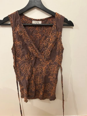 Brun omlottopp med orange broderi - Säljer en brun ärmlös omlottopp med orange blommigt broderi. Toppen har knytband i midjan och djup V-ringning framtill. 