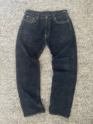 Mörkblå jeans från Evisu - Säljer ett par mörkblå jeans från Evisu med klassiska raka ben och kontrastsömmar. Baksidan har de ikoniska målade bågarna på bakfickorna och ett läderpatch i midjan. Jeansen är tillverkade i kraftigt raw denim och sitter ganska baggy, strl W 36 herr, bra skick med snygga slitningar vid benöppningarna