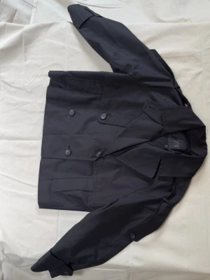 Svart kort trenchcoat från Zara - Svart kort något vattenavvisande trenchcoat från Zara med dubbelknäppning, hög krage och axelklaffar. Använd 1 gång. 
