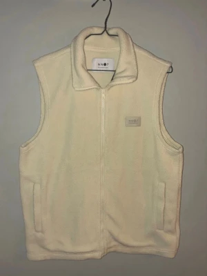 Beige fleeceväst från NN07 - Säljer en beige fleeceväst från NN07 med dragkedja framtill och två fickor. Västen har en hög krage och en diskret logotyp på bröstet. Perfekt för lager-på-lager och riktigt skön att bära under kyliga dagar.
