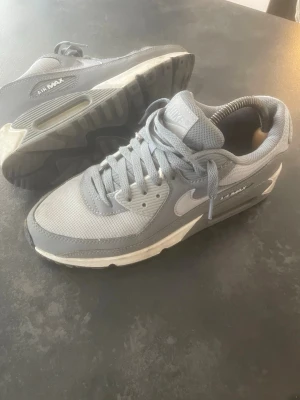Nike Air Max 90 grå sneakers - Snygga Nike Air Max 90 sneakers i olika grå nyanser med vit sula och klassisk Air Max-detalj på sidan. Ovandelen är i mesh och syntetmaterial för en sportig look. Ikonisk swoosh-logga på sidan och vadderad plös för extra komfort.