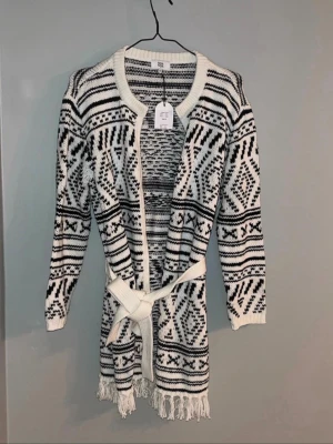 Mönstrad stickad kofta med knytband - Säljer en lång, stickad kofta i vitt och svart med grafiskt mönster. Koftan har ett knytband i midjan och fransar längst ner. Rund halsringning och långa ärmar. Perfekt för lager på lager och riktigt mysig.