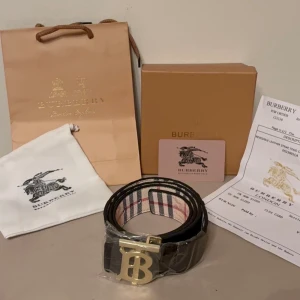 Burberry bälte med guldspänne - Snyggt Burberry bälte med klassiskt rutigt mönster på insidan och ett stort guldigt 'B'-spänne framtill. Bältet är svart på utsidan och har exklusiva detaljer som ger en lyxig känsla. Perfekt accessoar för att lyfta din outfit.