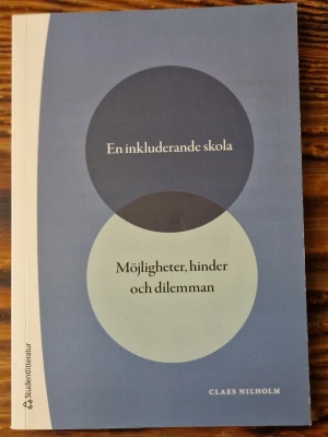 En inkluderande skola. Möjligheter, hinder och dilemman - Kursbok i pedagogik av Claes Nilholm med titeln 'En inkluderande skola: Möjligheter, hinder och dilemman'. Boken har ett blått och vitt omslag med grafiska cirklar och är tryckt på matt papper. Perfekt för dig som pluggar pedagogik eller specialpedagogik.