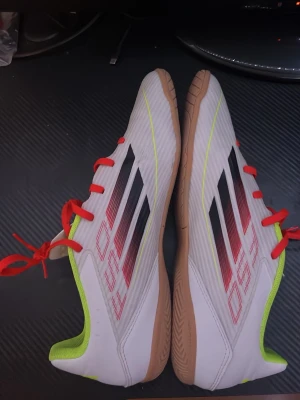 Adidas inneskor f50 - Säljer ett par grymma vita fotbollsskor från Adidas med röda snören och neon-gula insida. Skorna har klassiska svarta Adidas-stripes på sidan och en sula i ljusbrunt gummi, perfekt för inomhusspel. Materialet är syntetiskt och designen är slimmad och modern.