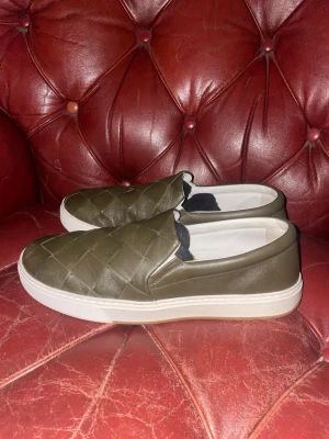 Olivgröna slip-on sneakers Bottega Veneta - Snygga olivgröna slip-on sneakers från Bottega Veneta i quiltat läder. Skorna har vit sula och diskret blå logga under sulan. Perfekta för dig som gillar stilrena och exklusiva detaljer. Klassisk låg modell som är enkel att matcha.