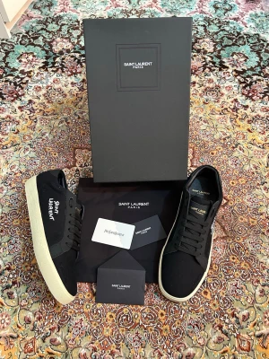 Svarta Saint Laurent sneakers - Snygga svarta sneakers från Saint Laurent med vit logga broderad på sidan och vit sula. Skorna har klassisk låg profil, svarta skosnören och är tillverkade i canvas med detaljer i läder. Perfekta för dig som gillar stilrena och exklusiva sneakers.