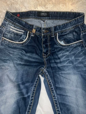 Blå jeans från SMOG, storlek 34/30 - Snygga blå jeans från SMOG med slitna detaljer och kontrastsömmar. Klassisk femficksmodell med vit kant på fickorna och markerade tvättade partier. Perfekta för en avslappnad och trendig look.