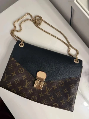 Brun Väska - Snygg handväska med klassiskt monogrammönster i brunt och guld. Väskan har svart lock i läder, guldfärgad kedjerem och elegant låsdetalj framtill. Röd på insidan❤️