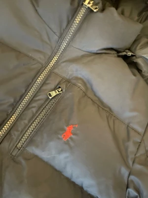 Mörkblå  pufferjacka från Polo Ralph Lauren - Säljer en mörkblå pufferjacka från Polo Ralph Lauren med röd broderad logga på bröstet. Jackan har dragkedja framtill, två sidofickor med dragkedja och en hög krage. Perfekt för kalla vinterdagar och riktigt snygg streetstil. Den ena fickan är lite sönder men går och laga. Skriv gärna prisförslag eller om du har några frågor❣️