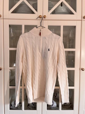 Vit kabelstickad tröja Polo Ralph Lauren - Vit kabelstickad tröja från Polo Ralph Lauren med halv dragkedja och ribbad hög krage. Tröjan har det klassiska broderade logotypen på bröstet och ribbade muddar vid ärmslut och nederkant. Perfekt för en stilren och avslappnad look.