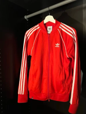 Röd Adidas träningsjacka med dragkedja - Klassisk röd Adidas träningsjacka med vita ränder längs ärmarna och Adidas-logga på bröstet. Jackan har hel dragkedja framtill, ribbade muddar och hög krage. Perfekt till träning eller sportig stil.