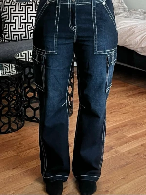 Mörkblå cargobyxor från True Religion - Säljer ett par mörkblå cargobyxor från True Religion med kontrasterande vita sömmar och flera stora fickor på sidorna. Byxorna har raka ben och hög midja, tillverkade i kraftigt denimtyg. Perfekta för en streetwear-look.