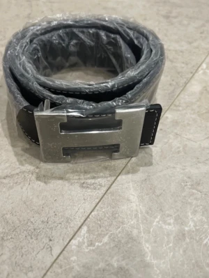 Svart bälte med silverfärgat H-spänne - Snyggt svart bälte med ett stort, silverfärgat H-format spänne. Bältet har vit kontrastsöm längs kanterna och är gjort i präglat skinn. Perfekt accessoar för att lyfta din outfit.