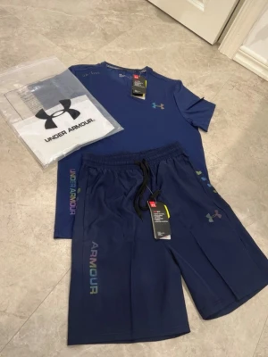 Blått träningsset från Under Armour - Snyggt blått träningsset från Under Armour med t-shirt och shorts. Båda plaggen har regnbågsfärgad logga och textdetaljer. Tillverkade i ett lätt och snabbtorkande material som håller dig sval under träningen. Perfekt för gymmet eller löprundan.