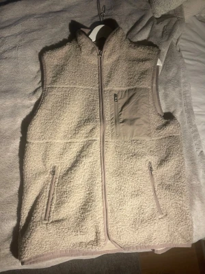 Beige teddyväst med dragkedja - Mysig beige teddyväst med hög krage och hel dragkedja framtill. Västen har två sidofickor med dragkedja och en bröstficka i kontrasterande tyg. Perfekt för lager-på-lager och chill stil. Materialet är fluffigt och ger en avslappnad vibe.