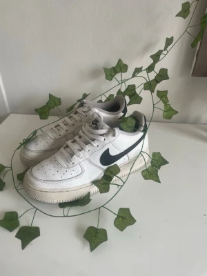 Nike vita Air Force 1 sneakers - Klassiska vita Nike Air Force 1 sneakers med svart swoosh på sidan. Skorna har lågt skaft, perforerad tåbox och tjock sula. Tillverkade i läder med textildetaljer och har en retro vibe som passar till streetwear och casual outfits.