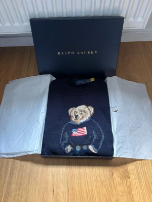 Stickad Polo Ralph lauren bear - Mörkblå stickad tröja från Polo Ralph Lauren med ikonisk björn på bröstet som bär en tröja med amerikanska flaggan. Tröjan är i absolut topp skick i storlek m men passar s hör av er vid frågor eller funderingar priset går att diskutera!!