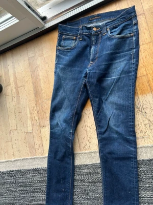 Blå jeans från Nudie Jeans - Säljer ett par klassiska blå jeans från Nudie Jeans med snygga slitningar och kontrastsömmar. Jeansen har fem fickor, normal midja och raka ben. Bakfickorna har Nudie Jeans ikoniska broderade vågmönster. Perfekta till en avslappnad stil. W 31 passar w29-28. Väldigt korta 