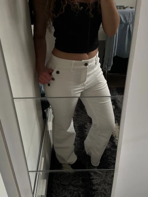Vita jeans med knappar i sidan - Säljer ett par vita jeans med raka ben och snygga dekorativa knappar längs sidan vid höften. Jeansen har hög midja och klassiska fickor både fram och bak. Perfekta för dig som vill sticka ut lite extra med detaljer. Materialet känns mjukt och stretchigt.