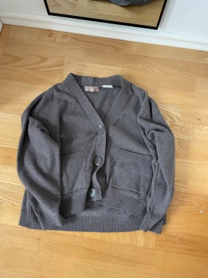 Grå kofta med stora knappar - Mysig grå kofta med v-ringning och tre stora knappar framtill. Två rymliga fickor på magen och ribbade muddar vid ärmslut och nederkant. 
