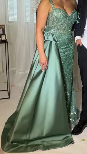 Grön långklänning med paljettdetaljer - Elegant grön långklänning med glittrande paljetter upptill och på ena sidan. Klänningen har en vacker släp och en axelbandslös design med markerad midja och satinkänsla i tyget. Perfekt för speciella tillfällen.
