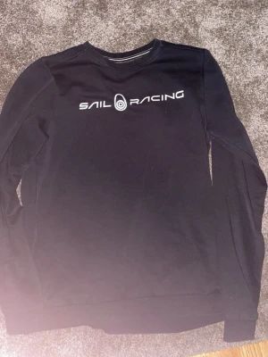 Svart tröja från Sail Racing - Svart långärmad tröja från Sail Racing med vit logga och text på bröstet. Klassisk rund halsringning och mjukt material som känns skönt mot huden. Perfekt för en clean och sportig look.
