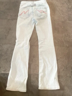 Ljusblå bootcut jeans med slitningar - Säljer ett par ljusblå bootcut jeans med slitna hål på knäna och broderade detaljer i rosa på bakfickorna. Jeansen har klassisk femficksdesign och är tillverkade i denim. Perfekta för en avslappnad och trendig look.