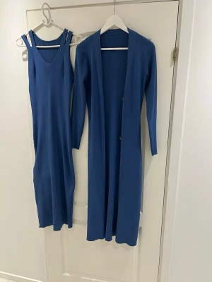 Blå ribbad långklänning med kofta - Säljer en blå ribbad långklänning med matchande lång kofta. Klänningen är ärmlös med v-ringning och koftan har långa ärmar samt knappar framtill. Perfekt för dig som gillar en stilren och bekväm look.